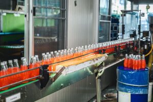 Bottles filled with orange soda move along a conveyor belt in a beverage factory production line, passing through an automatic label applicator for precise labeling.